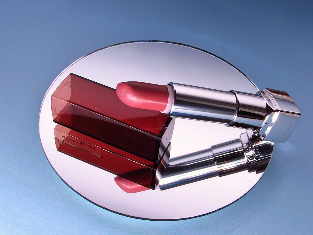 Maybelline Velvet Beige lipstick case and a cap on a mirror surface with blue background